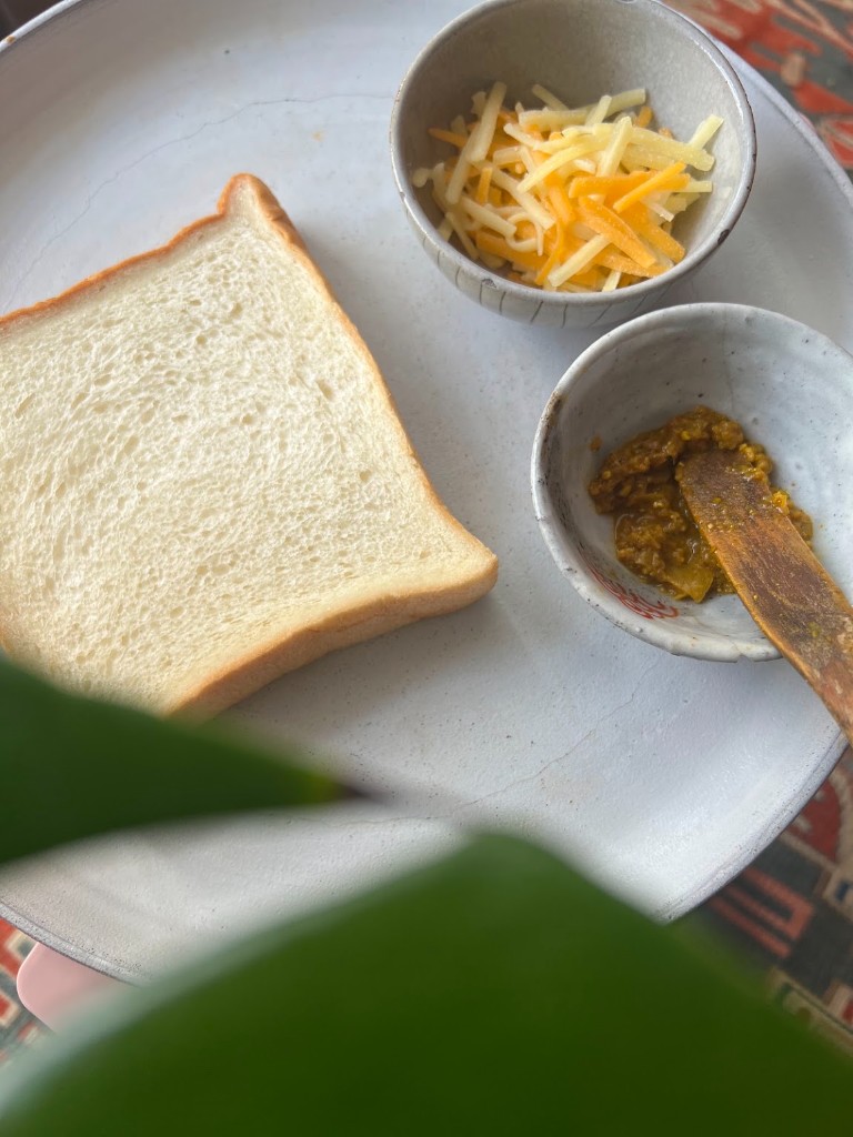 Bahan roti panggang keju Curry Koji: sepotong roti tawar tebal, keju parut, dan Curry Koji di atas piring keramik.